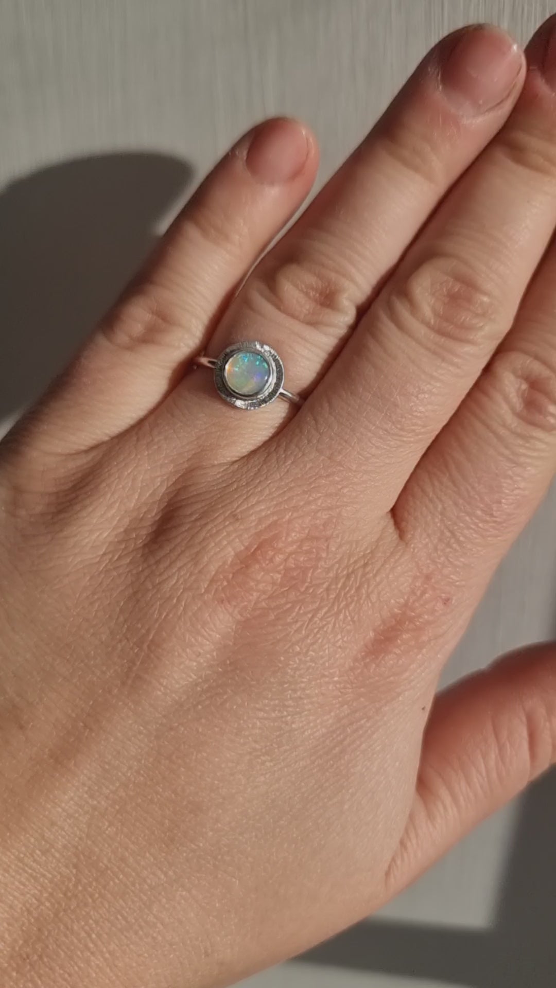 A handmade sterling silver ring featuring an Australian Crystal Opal from Coober Pedy. The opal features pinks, greens, and blue swirls of colour and is set in a radiant engraved silver setting. It is being worn by a hand and is showing off the play of colours in the sun.